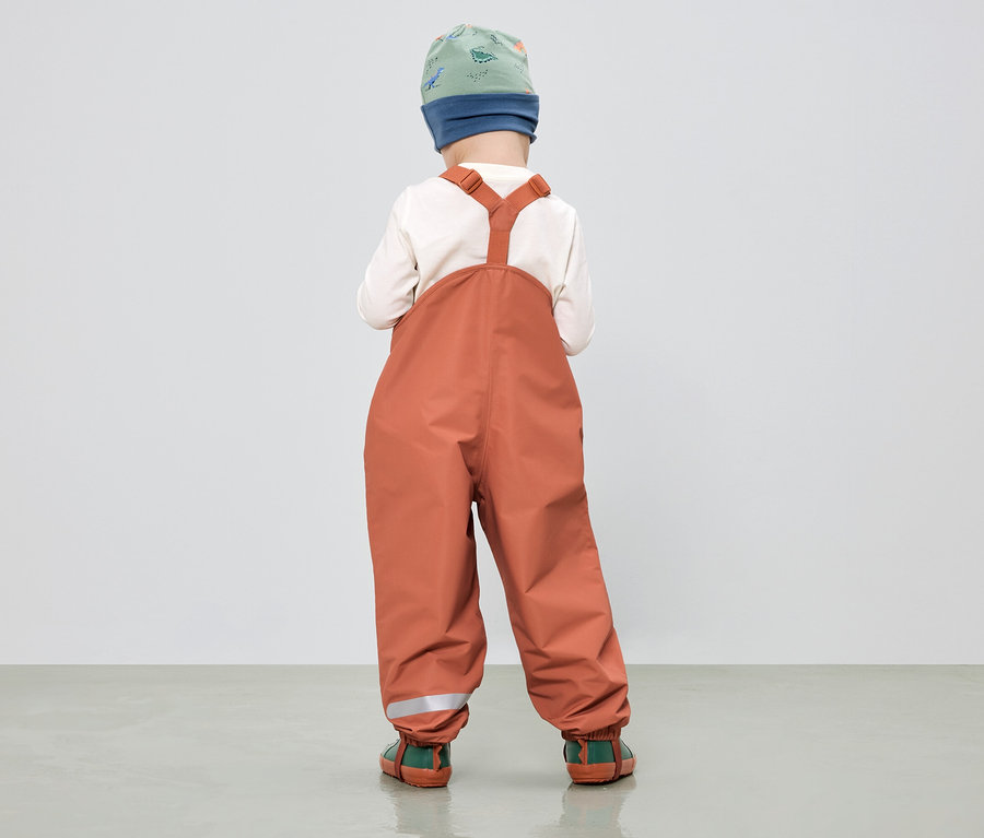 Kind von hinten in orangefarbener Matschhose mit Hosenträgern, grün-blau gemusterter Mütze und cremefarbenem Oberteil.
