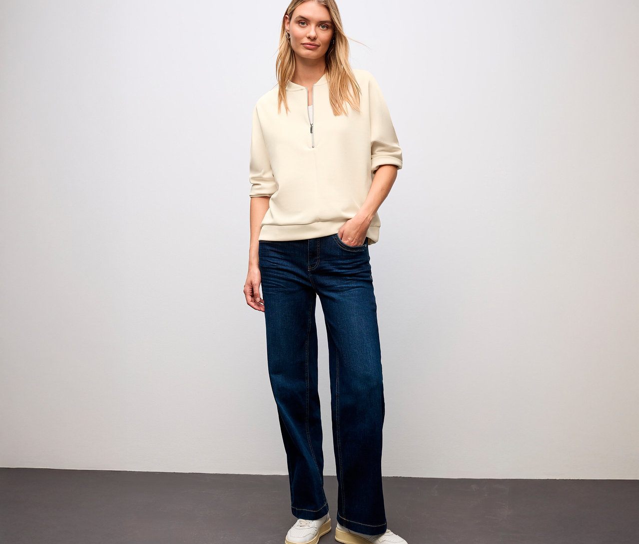 Eine blonde Frau steht und trägt ein Street One T-Shirt aus Modalmix, Jeans und Turnschuhe.