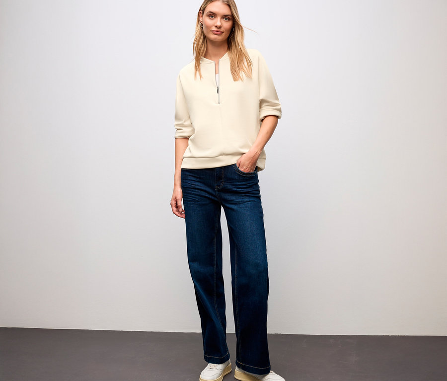Eine blonde Frau steht und trägt ein Street One T-Shirt aus Modalmix, Jeans und Turnschuhe.