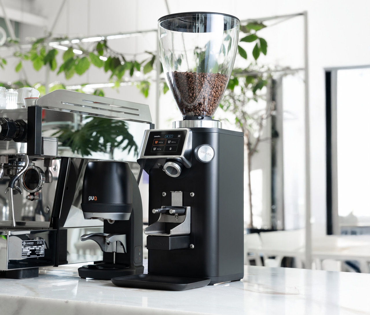 Auf einer Marmoroberfläche stehen eine Kaffeemühle mit Bohnen im Trichter, eine Espressomaschine und ein Pucks Screen.