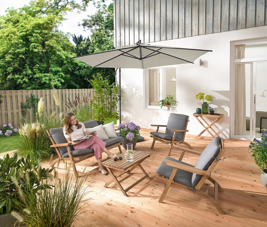 Eine Frau sitzt auf einem zusammenklappbaren Garten-Lounge-Set mit Premium-Kissen, liest und ist umgeben von einem Garten-Tabletttisch und 2 Outdoor-Kissen mit Leinen-Haptik.