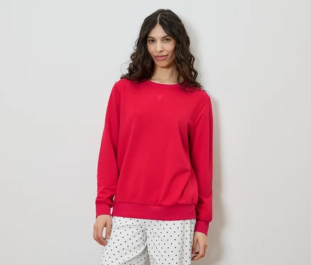 Eine Frau mit lockigem, braunem Haar trägt einen roten Pullover und eine gepunktete Hose vor einem weißen Hintergrund.