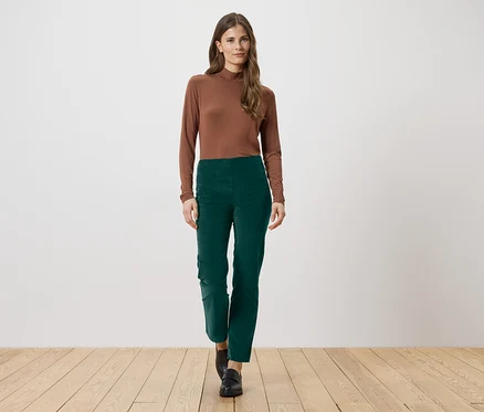 Eine Frau mit langen braunen Haaren trägt ein braunes Langarmshirt und eine grüne Hose. Sie steht auf einem Holzboden.