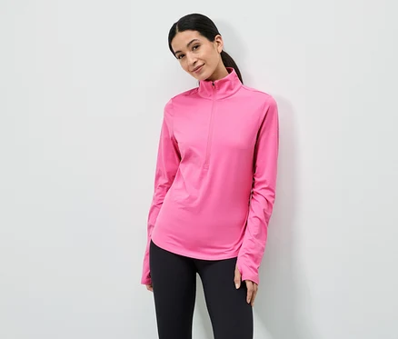 Eine Frau mit Pferdeschwanz trägt ein pinkes, langärmeliges Oberteil mit Reißverschluss und schwarze Leggings vor einer weißen Wand.