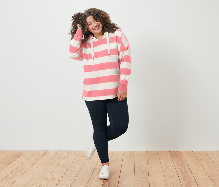 Frau mit lockigem Haar trägt einen gestreiften Hoodie und Leggings und lächelt.