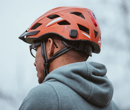 Nahaufnahme eines Mannes mit Brille und grauem Hoodie, der einen orangefarbenen FISCHER Fahrradhelm Urban PLUS Miami trägt.