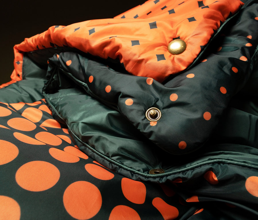 Nahaufnahme einer gefalteten, wendbaren Daunenjacke in Grün mit orangefarbenen Punkten und Orange mit schwarzen Quadraten.