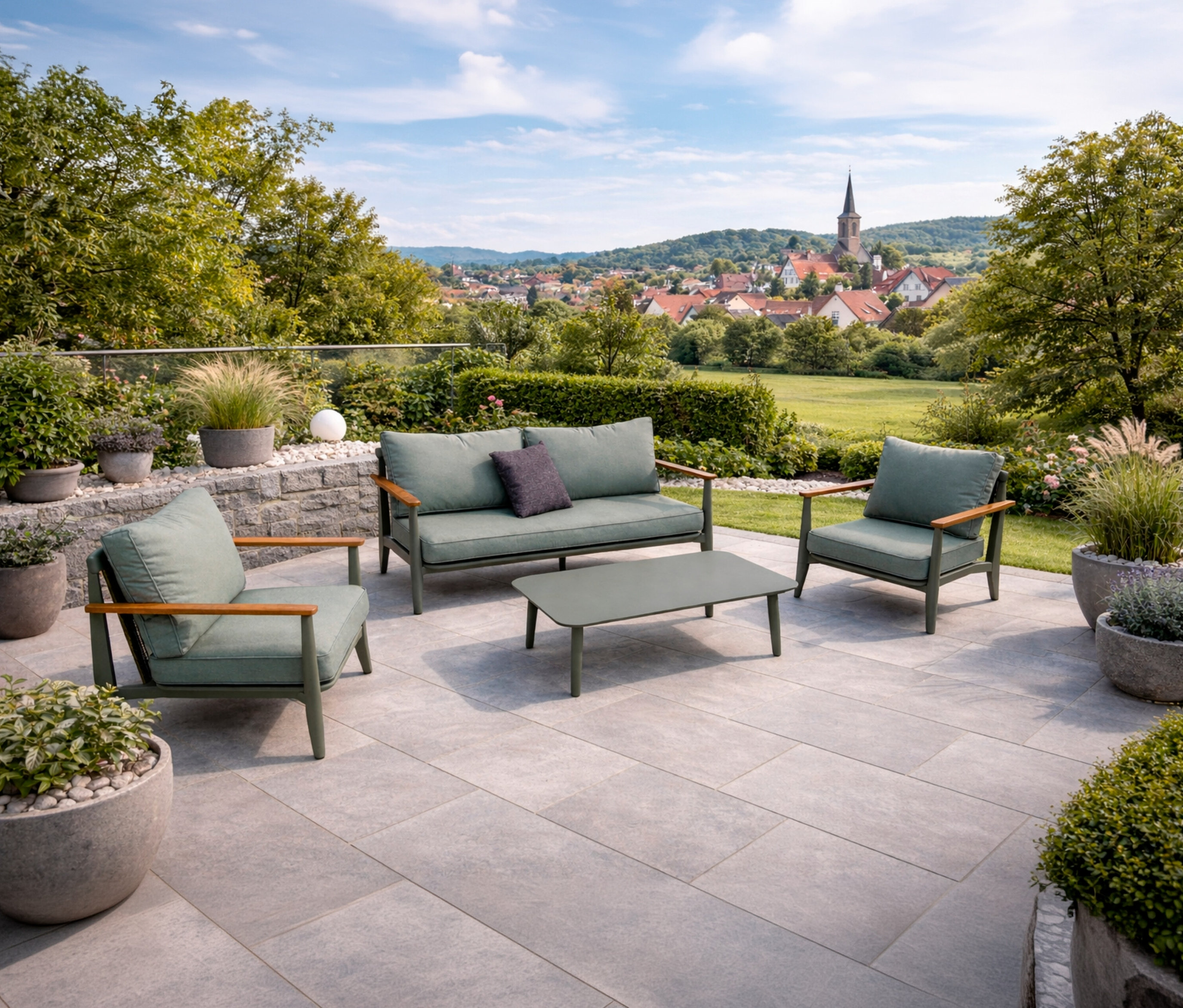 Auf einer Terrasse steht ein Leco Lounge-Set inkl. Tisch mit grünen Polstern und Holzarmlehnen vor einer Landschaft mit Häusern und Bäumen.