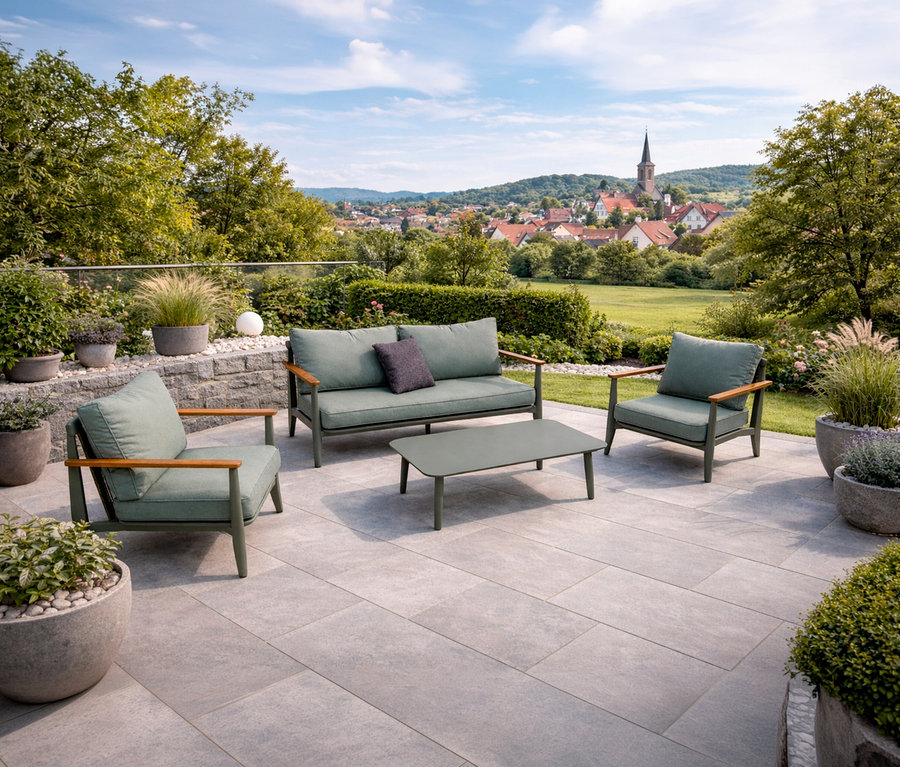 Auf einer Terrasse steht ein Leco Lounge-Set inkl. Tisch mit grünen Polstern und Holzarmlehnen vor einer Landschaft mit Häusern und Bäumen.
