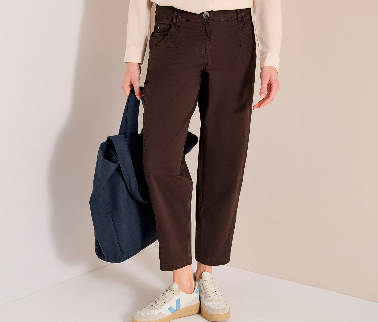 Eine Person trägt ein cremefarbenes Oberteil, eine braune CECIL Barrel »Scarlett« Hose und helle Sneaker mit blauen Akzenten, während sie eine dunkelblaue Stofftasche hält.