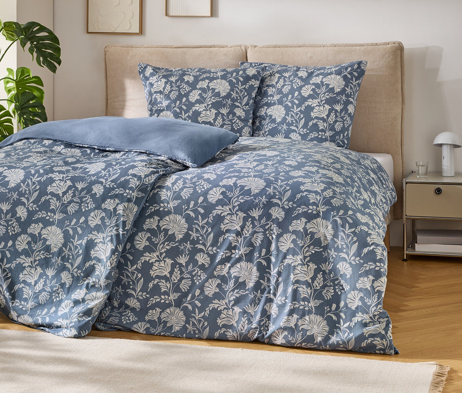 Ein Bett mit blauer Bettwäsche mit weißem Blumenmuster und einem hellblauen Kissen. Das Bett hat ein beiges Kopfteil. Links steht eine Monstera-Pflanze, rechts ein Nachttisch mit Lampe und Glas. Ein beiger Teppich liegt auf dem Holzboden.