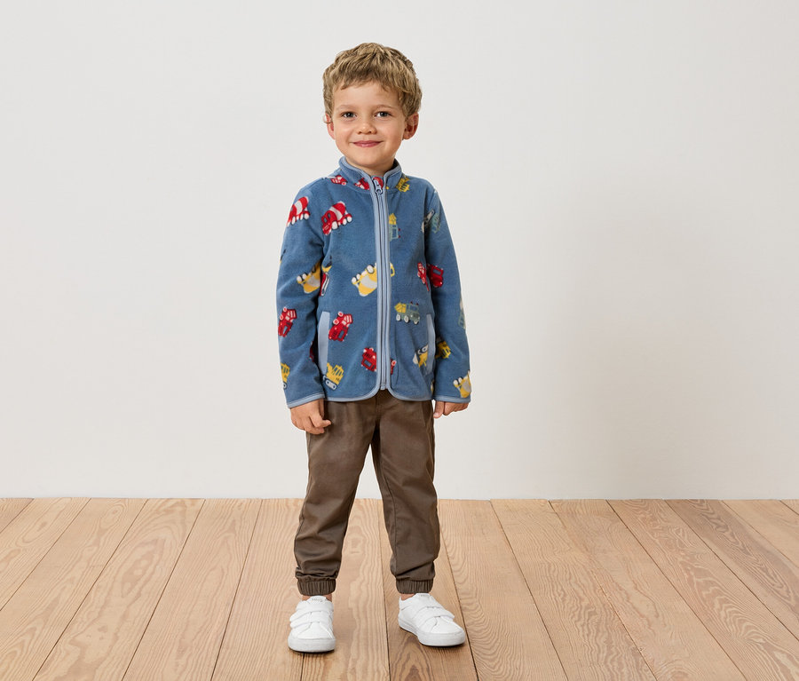 Ein Junge steht in einer blauen Fleecejacke mit Fahrzeugmuster, braunen Hosen und weißen Schuhen vor einer Wand.