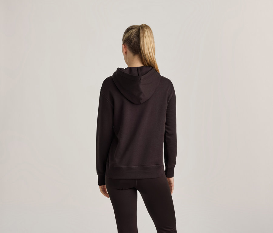 Frau von hinten, trägt einen dunklen Kapuzenpullover und dunkle Leggings. Ihr blondes Haar ist zu einem hohen Pferdeschwanz gebunden.