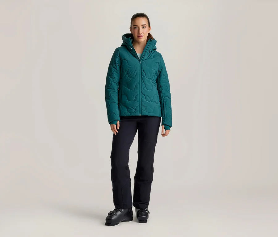 Frau steht in einer türkisfarbenen Steppjacke mit Kapuze, schwarzen Skihosen und schwarzen Skischuhen.