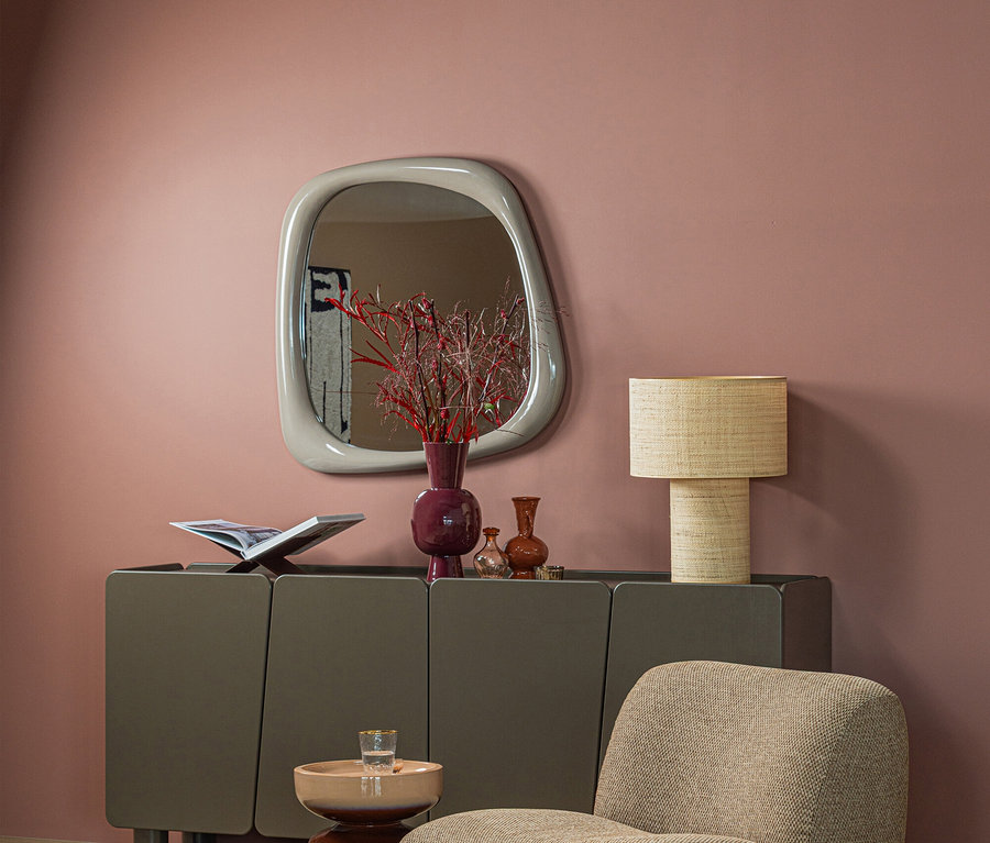 Modernes Wohnzimmer mit rosa Wand. Ein dunkelgraues Sideboard steht unter einem unregelmäßig geformten Spiegel. Darauf sind ein offenes Buch, eine lila Vase mit roten Zweigen, kleinere Vasen und eine geflochtene Lampe. Davor stehen ein beiger Sessel und ein kleiner Beistelltisch auf einem braunen Teppich.