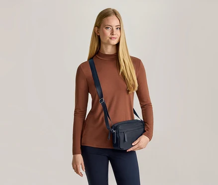 Eine Frau mit langen blonden Haaren trägt ein braunes Langarmshirt, eine dunkelblaue Hose und eine dunkelblaue Umhängetasche. Sie hält die Tasche mit ihrer linken Hand und blickt nach vorne.