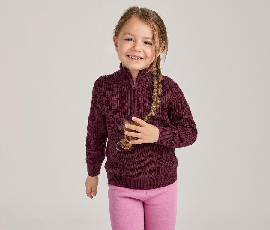 Lächelndes junges Mädchen mit langem Zopf, das einen burgunderfarbenen gerippten Half-Zip-Pullover und pinke Leggings trägt und ihren Zopf mit der rechten Hand hält.