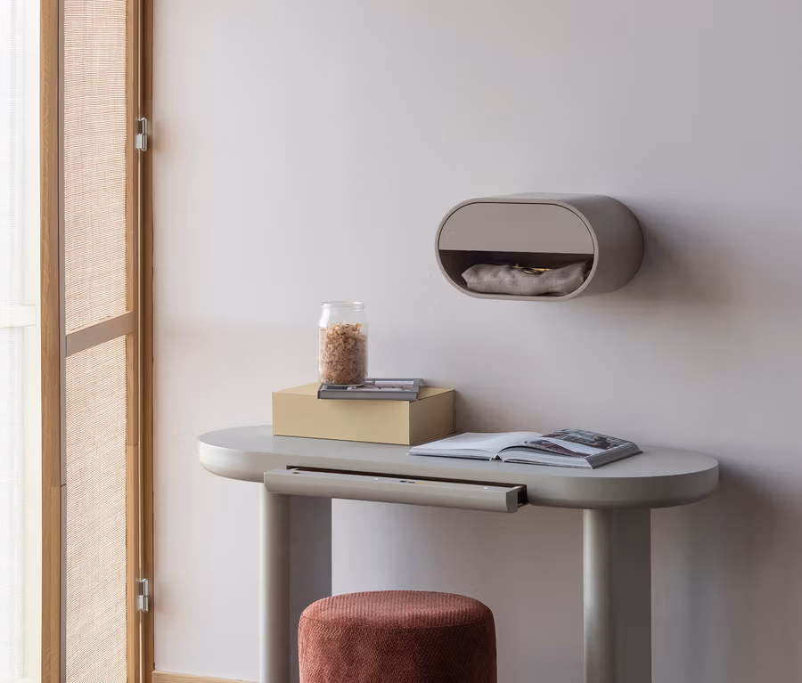 Ein minimalistischer Raum mit einem hellgrauen Konsolentisch, einem rostfarbenen Samthocker und einem ovalen Wandschrank. Auf dem Tisch stehen eine gelbe Schachtel, ein Glasgefäß und ein offenes Buch. Links ist ein geflochtener Paravent teilweise geöffnet.