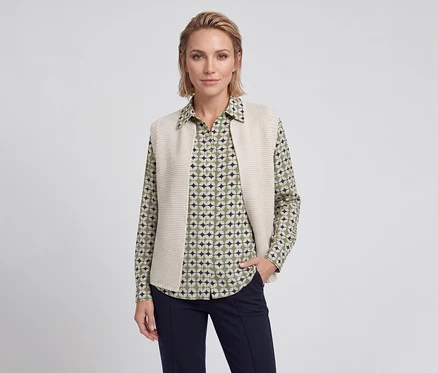 Frau mit kurzen blonden Haaren trägt ein grün, beige und dunkelblau gemustertes Hemd, eine beige Strickweste und eine dunkelblaue Hose, eine Hand in der Tasche.