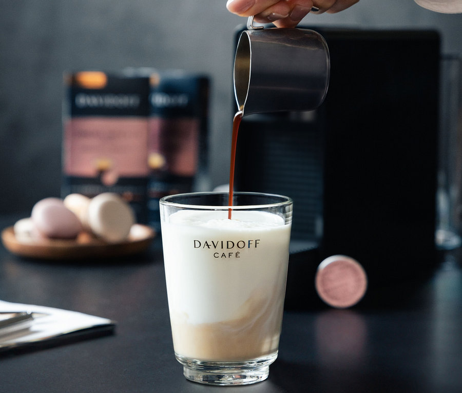 Eine Hand gießt Kaffee in ein Glas Milch. Im Hintergrund befinden sich eine Kaffeemaschine und eine Packung DAVIDOFF CAFÉ Crema Intense.