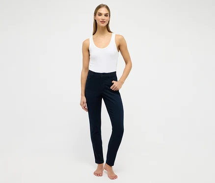 Frau steht, trägt ein weißes geripptes Tanktop und dunkelblaue High-Waist-Jeans, die linke Hand in der Tasche. Sie ist barfuß.