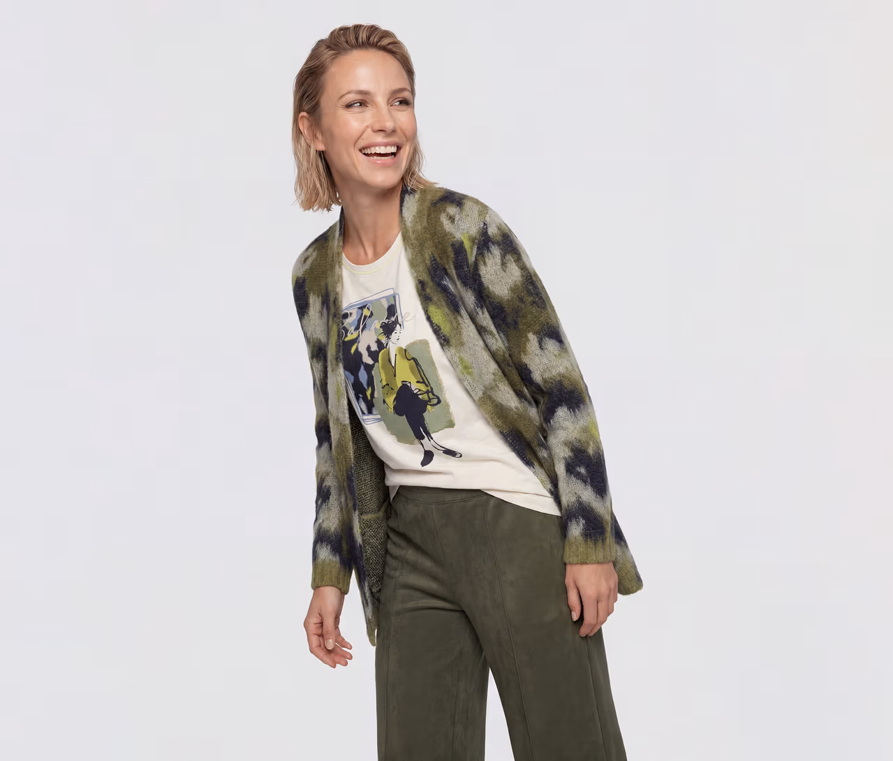 Eine lächelnde Frau mit kurzen blonden Haaren blickt nach rechts, trägt eine grün-schwarz gemusterte Strickjacke über einem cremefarbenen T-Shirt mit Grafikdruck und olivgrüne Hosen.