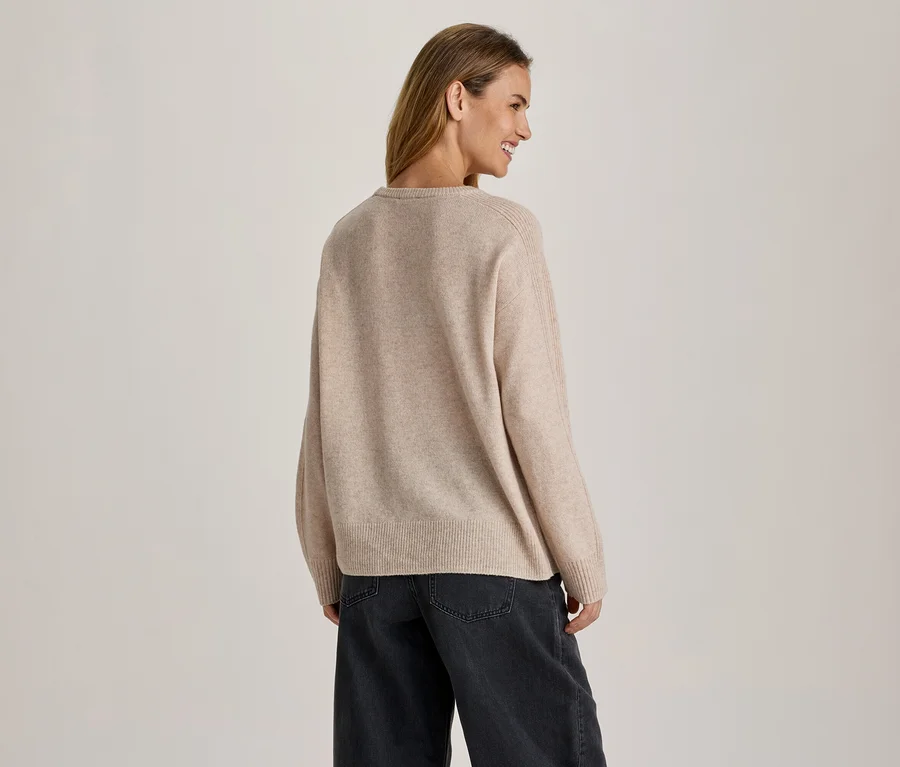 Frau von hinten, leicht nach rechts gedreht und lächelnd, trägt einen beigen Pullover und eine dunkle weite Hose.