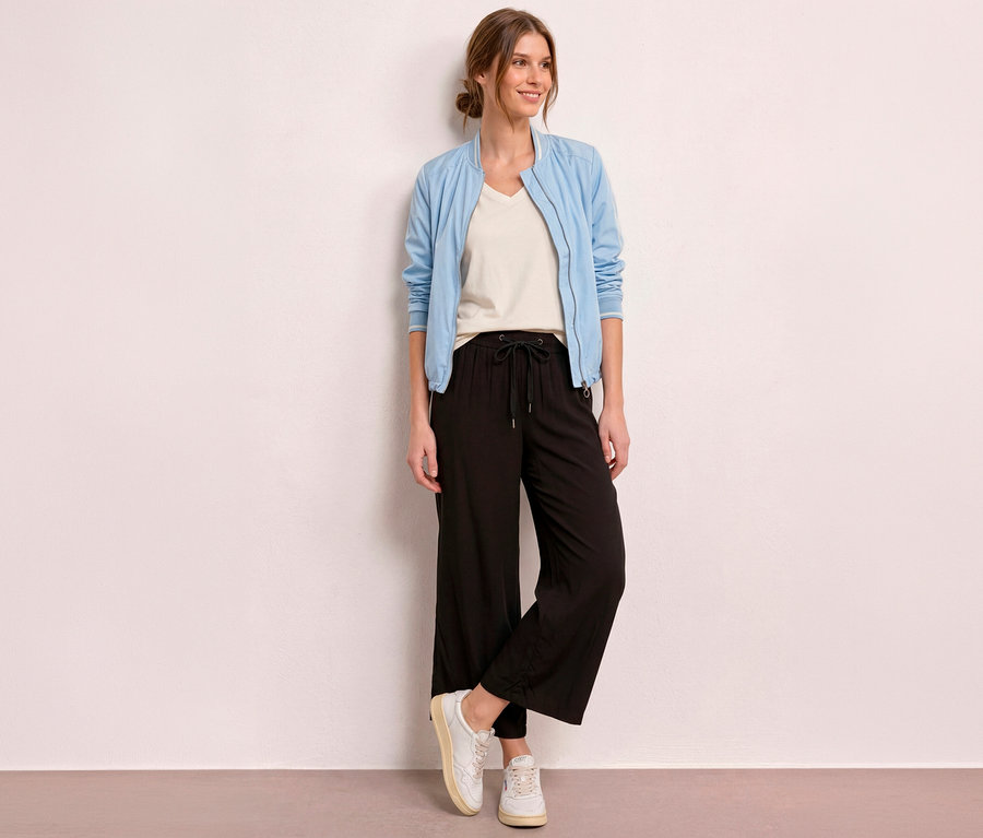 Eine lächelnde Frau steht vor einer hellen Wand und trägt eine hellblaue Bomberjacke, ein cremefarbenes T-Shirt und eine schwarze Street One 7/8-Culotte »EMMA«.