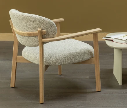 Heller Holz-Sessel mit hellgrauem, strukturiertem Bezug und Armlehnen, daneben ein beiger Beistelltisch mit offenem Buch. Hintergrund ist eine senfgelbe Wand und dunkler Holzboden.