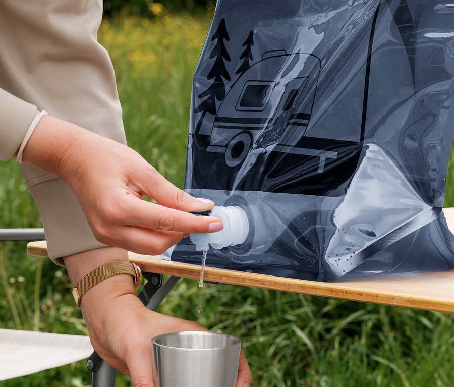 Eine Person füllt Wasser aus einem dunklen, flexiblen faltbaren Wasserkanister mit Camper-Motiv in einen silbernen Becher. Der Kanister steht auf einem Holztisch im Freien.