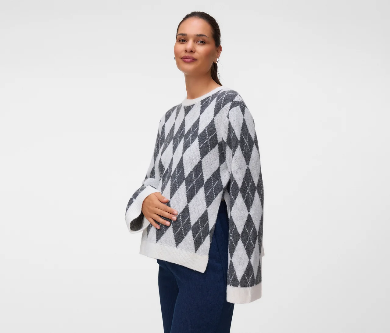 Frau in einem grau-weißen Rautenpullover mit Seitenschlitzen und dunkelblauer Hose, blickt in die Kamera und hält eine Hand auf ihren Bauch.