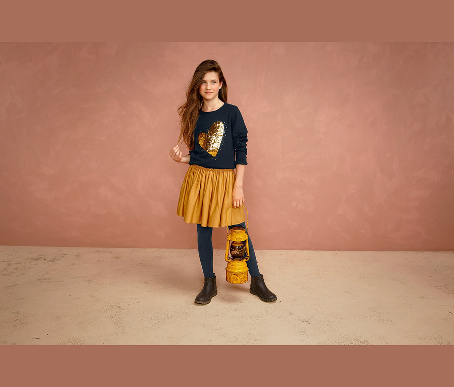 Mädchen steht mit gelber Laterne, trägt marineblauen Pullover mit goldenem Herz und curryfarbenen Rock.