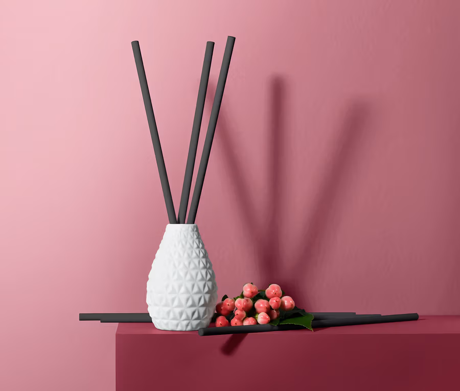 Raumduft-Set »Zedernholz« mit weisser Vase und schwarzen Stäbchen auf weinrotem Sockel vor rosa Hintergrund.