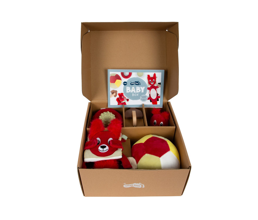 Offene braune Babybox mit mehreren Fächern, gefüllt mit roten Plüschtieren, einem rot-gelben Ball, einem grünen Beißring, zwei Holzpilzen und einem "BABY BOX"-Heft.
