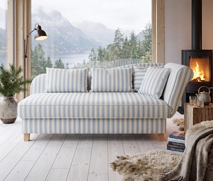 Ein gemütliches Wohnzimmer mit einem hellblau-weiß gestreiften Sofa vor einem großen Fenster mit Blick auf eine bergige Fjordlandschaft. Links vom Sofa stehen eine Stehlampe und eine Topfpflanze. Rechts davon befinden sich ein schwarzer Holzofen mit Feuer und ein Holztischchen mit einer Teekanne und Gläsern. Ein Kunstfellteppich, Bücher und eine Decke liegen auf dem hellen Holzboden.