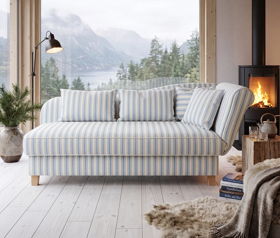 Ein gemütliches Wohnzimmer mit einem hellblau-weiß gestreiften Sofa vor einem großen Fenster mit Blick auf eine bergige Fjordlandschaft. Links vom Sofa stehen eine Stehlampe und eine Topfpflanze. Rechts davon befinden sich ein schwarzer Holzofen mit Feuer und ein Holztischchen mit einer Teekanne und Gläsern. Ein Kunstfellteppich, Bücher und eine Decke liegen auf dem hellen Holzboden.