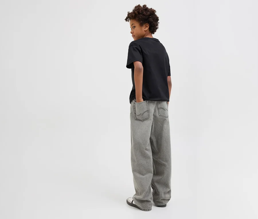 Junge von hinten in schwarzem T-Shirt und grauen Jack&Jones JJOrginial Alex Jeans, Hände in Hosentaschen, blickt über rechte Schulter.