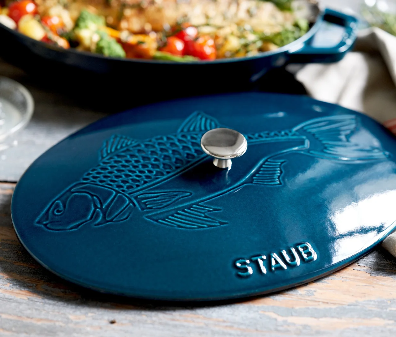 Dunkelblauer, ovaler Staub-Deckel mit geprägtem Fischmotiv und silbernem Knauf auf einer Holzoberfläche. Im unscharfen Hintergrund ist eine Auflaufform mit Essen zu sehen.