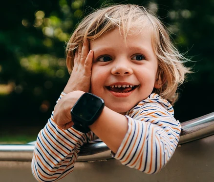 Ein lächelndes Kind mit blondem Haar und gestreiftem Shirt trägt eine schwarze Xplora XGO3 Kids Watch am Handgelenk.