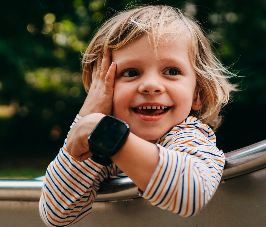 Ein lächelndes Kind mit blondem Haar und gestreiftem Shirt trägt eine schwarze Xplora XGO3 Kids Watch am Handgelenk.
