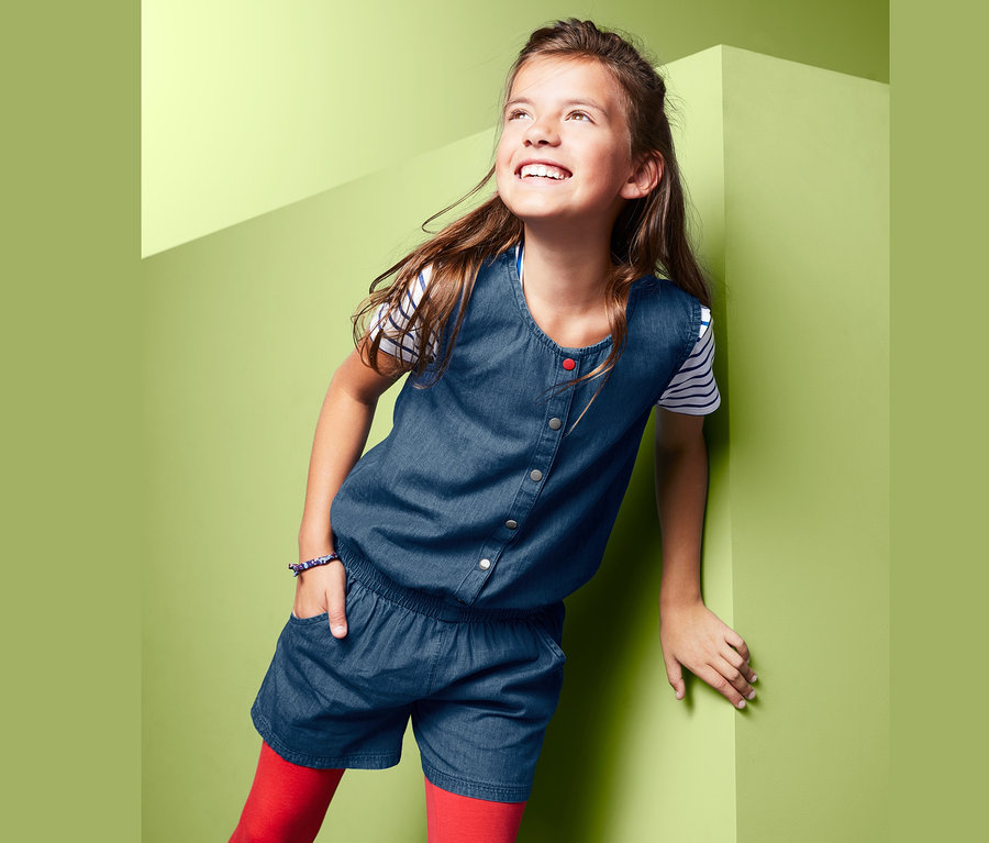 Lächelndes Mädchen in kurzem Jumpsuit in leichter Denim-Qualität lehnt an einer grünen Wand.