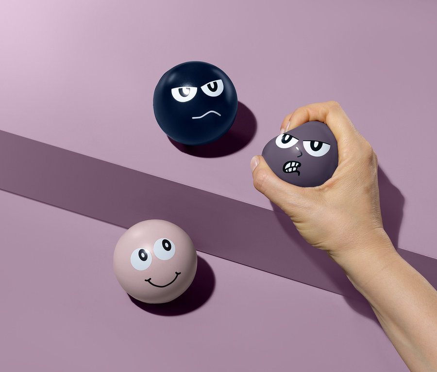 Eine Hand drückt einen lila Antistressball mit einem Stirnrunzeln. Auf dem lila Hintergrund liegen zwei weitere Antistressbälle.