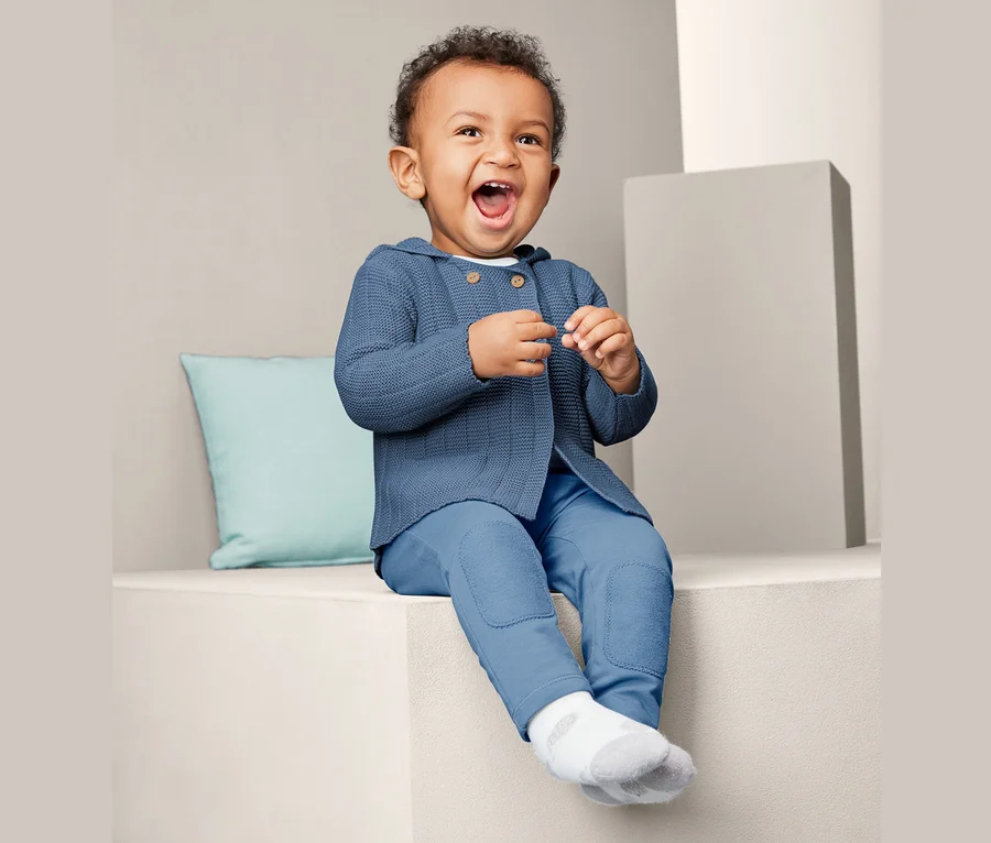 Lächelndes Baby sitzt und trägt eine blaue Strickjacke.