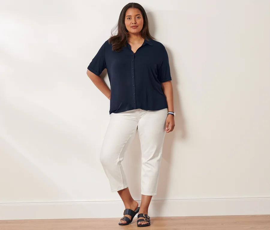 Frau posiert in Blusenshirt mit Knopfleiste, Slimfit-Jeans und Sandalen mit Schnalle. Sie trägt eine Quarz-Armbanduhr aus Edelstahl.