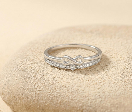 Silberner Amor Ring mit Infinity-Symbol und glitzernden Steinen auf einer sandfarbenen Oberfläche.