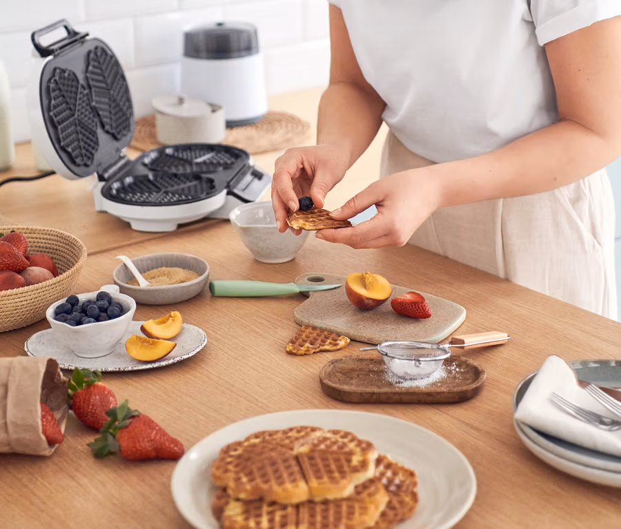 Frau dekoriert Waffeln mit Blaubeeren an einem Tisch mit Waffeln, einer Eismaschine »Comfortice« und dem WILFA Waffeleisen »TRADITION DOUBLE WDWA-516W«.