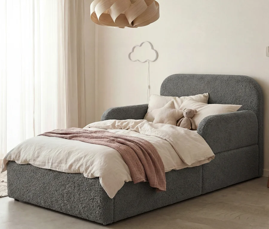 Ein graues Bouclé-Bett mit Kopf- und Armlehnen, bedeckt mit einer hellen Bettdecke und einer rosa Decke. Mehrere Kissen und ein Teddybär liegen am Kopfende. Eine Wolken-Wandleuchte und eine Holz-Pendelleuchte sind sichtbar.