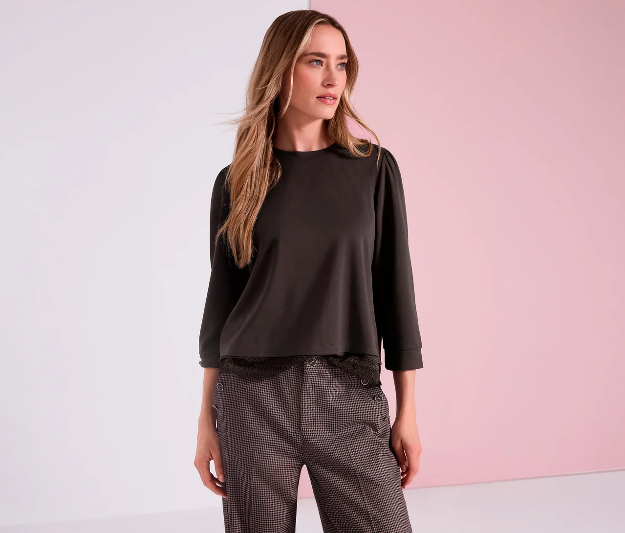 Eine Frau trägt ein braunes Street One 3/4-Jerseyshirt mit Puffärmeln und eine gemusterte Hose vor einem rosa-weißen Hintergrund.