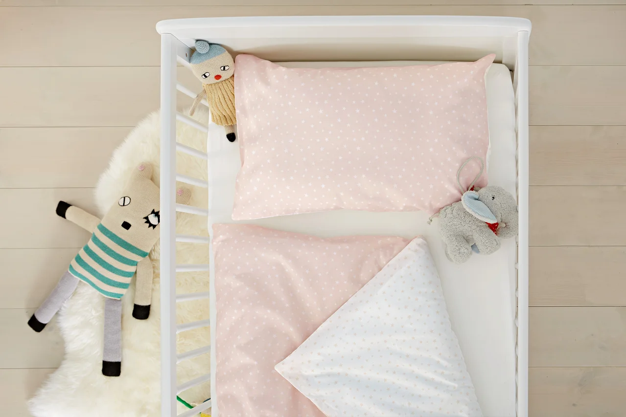 Draufsicht auf ein weißes Kinderbett mit rosa Bettwäsche mit Sternen, einem Stofftier und einer Decke.