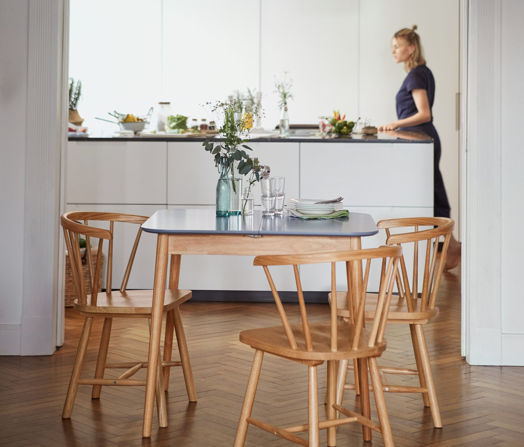 Ausziehbarer Küchentisch (ca. 90–120 x 80 cm) mit vier Holzstühlen im Vordergrund, im Hintergrund eine Frau in der Küche.